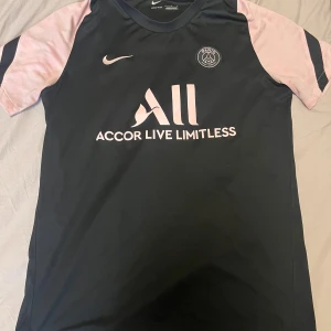 PSG fotbollströja Nike svart/rosa - Paris Saint-Germain fotbollströja från Nike med svart kropp och ljusrosa korta ärmar. PSG-logga och Nike Swoosh på bröstet, samt sponsortryck 'Accor Live Limitless' och 'Visit Rwanda' i rosa. Tillverkad i lätt, ventilerande material perfekt för träning.