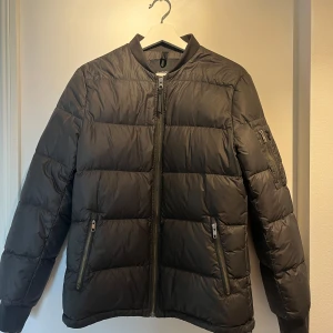 Svart pufferjacka med dragkedjor - Säljer en svart pufferjacka med quiltad design och ribbade muddar. 