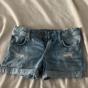 Ljusblå jeansshorts från H&M &Denim - Snygga ljusblå jeansshorts från H&M &Denim med slitna detaljer och uppvikta benslut. Klassisk femficksmodell med knapp och dragkedja framtill. Perfekt för chill dagar och sommarhäng.