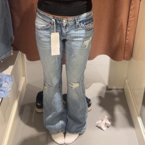 Ljusblå bootcut jeans med röda detaljer och hål - Snygga ljusblå jeans med bootcut passform och slitna detaljer på låren och knäna. Jeansen har klassiska fem fickor, markerade sömmar och knäppning fram. Perfekta för dig som gillar en avslappnad och trendig look.