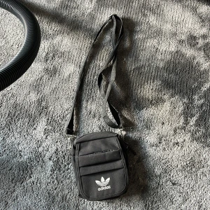 Svart beknarväska från Adidas - Svart beknarväska från Adidas med klassisk vit logga framtill. Väskan har justerbar rem, dragkedja och flera fack för småprylar. Tillverkad i slitstark polyester och perfekt för att bära med sig det viktigaste. Enkel och stilren design som funkar till det mesta.