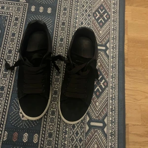 Svarta sneakers från Alexander McQueen - Stilrena svarta sneakers från Alexander McQueen med vit sula och guldfärgad logga på hälen. Skorna har rund tå, breda svarta skosnören och är tillverkade i mocka. Perfekta för dig som gillar en clean och lyxig look.