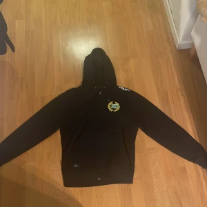 Svart Hammarby ziphoodie från Craft - Svart ziphoodie från Craft med Hammarby IF-logga på bröstet. Tröjan har huva med snörning, dragkedja framtill och långa ärmar. Perfekt för dig som vill visa ditt stöd för Hammarby och samtidigt hålla dig bekväm och snygg. Tröjan är i storlek Xs men passar även S bra.