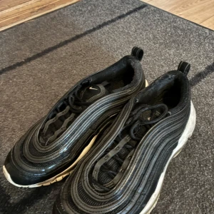 Nike Air Max 97 svart/vit sneakers - Säljer ett par Nike Air Max 97 i svart med vita detaljer. Skorna har den ikoniska vågiga designen, snörning och en synlig Air-sula. Perfekta för dig som gillar streetstyle och vill ha en klassisk sneaker med modern vibe.