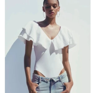 Vit volangbody med v-ringning - Superfin vit body med djup v-ringning och stora volanger över axlarna. Toppen är ärmlös och har en snygg öppen rygg. Perfekt för en trendig och fräsch look till sommaren.