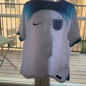 England tröja - England fotbollströja från Nike med Maguire och nummer 6 på ryggen. Tröjan är vit med blå detaljer och har det klassiska landslagsmärket på bröstet. Tillverkad i Dri-FIT-material som andas. Kortärmad modell med blå och turkosa inslag vid axlar och ärmslut.