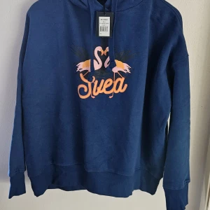 Blå hoodie från Svea med flamingos - Blå hoodie från Svea i modellen Flamingo Hood, med tryck av två flamingos och Svea-logga i rosa och orange på bröstet. Tröjan har huva, långa ärmar och är gjord i mjuk bomull och polyester. Perfekt för chill dagar och streetstyle.