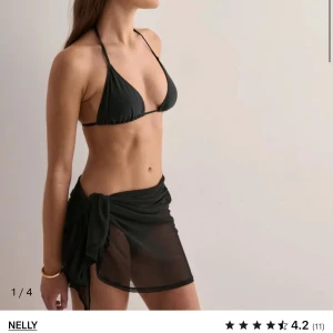 Svart kjol med sarong från Nelly - Snygg svart kjol från Nelly med trekantsöverdel och knyt i nacken. Matchas med en transparent svart sarong som knyts i sidan. Perfekt för stranden och ger en stilren look. Tillverkad i mjukt syntetmaterial som torkar snabbt.