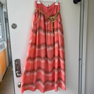 Lång randig klänning med knytband - Säljer en lång klänning i korallrosa med vågiga ränder i beige och brunt. Klänningen har axelbandslös design och ett snyggt gult knytband med ring i midjan. Materialet känns lätt och flowy, perfekt för varma dagar. Fickor på sidorna för extra chill vibe.