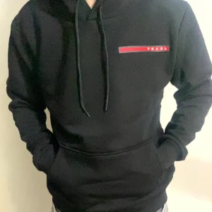Svart hoodie från Prada med logga - Svart hoodie från Prada med klassisk huva och snörning. Diskret röd och vit logga på bröstet. Mjuk och skön passform, perfekt för en chill och stilren look.