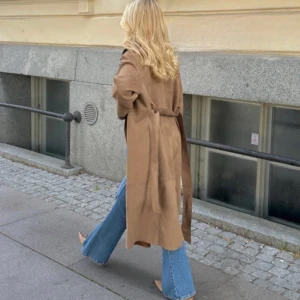 Brun trenchcoat i mocka-look - Snygg brun trenchcoat med bälte i midjan och långa ärmar. Jackan har en klassisk, rak passform och är tillverkad i ett material som ser ut som mocka. Perfekt för lager-på-lager och ger en chill men stilren vibe till din outfit. Har tyvärr tappat bandet men lika snygg ändå! 