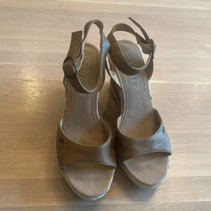Bruna kilklackade sandaler från Esprit - Snygga bruna sandaler från Esprit med kilklack och flätad detalj runt sulan. Skorna har öppen tå, justerbart spänne runt vristen och ovandel i skinn. Perfekta för varma dagar när du vill ha både stil och komfort. Knappt använda. Storlek 39