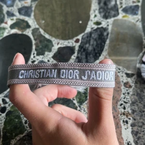 Grå Christian Dior J'Adior armband - Snyggt grått armband från Christian Dior med texten 'J'Adior' broderad i vitt. Armbandet har en bred, vävd design och avslutas med två stilrena tofsar. Perfekt accessoar för att lyfta din outfit med en lyxig touch.