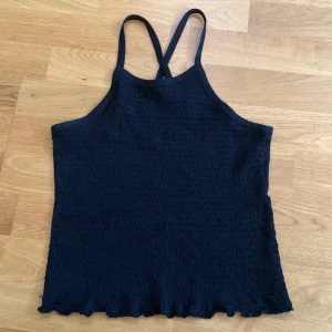 Svart smockad linnetopp från Lindex - Svart linnetopp från Lindex med smockad struktur och tunna axelband som korsas i ryggen. Toppen har en lätt vågig kant nedtill och är ärmlös och stretchig. Du står för frakten, vid mer info skriv
