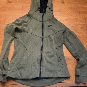 Olivgrön Nike tech med huva M - Snygg olivgrön flis från Nike med svart logga på bröstet. Jackan har hel dragkedja, justerbar huva och två fickor framtill. Tillverkad i mjuk polyesterblandning, perfekt för chilliga dagar. Tumhål i ärmsluten för extra komfort.
