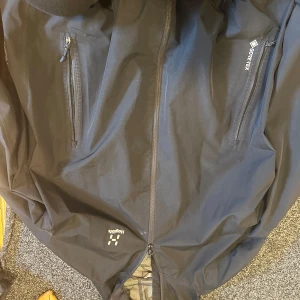 Svart Gore-Tex vindjacka från Haglöfs - Svart vindjacka från Haglöfs i slitstark Gore-Tex med dragkedja framtill och två vertikala fickor med dragkedja. Jackan har en sportig look och är perfekt för uteliv. Klassisk passform och diskret logga på bröstet.