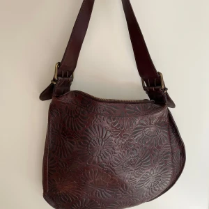 Brun axelväska i skinn från Fendi - Säljer en brun oyster bag från Fendi i äkta skinn med präglat blommönster. Väskan har justerbar axelrem med spännen i mässing och stängs med dragkedja. Insidan är fodrad i beige tyg och har en metallbricka med Fendi-logga.