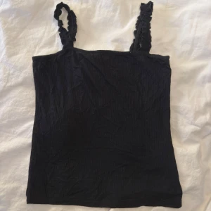 Svart ribbad linnetopp med volangband - Snygg svart linnetopp med ribbad struktur och volangprydda axelband. Perfekt att styla med jeans eller kjol för en trendig look. Axelbandsmodellen ger en somrig vibe och den svarta färgen gör den lätt att matcha.