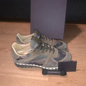 Valentino Rockrunner sneakers  - Snygga Valentino Rockrunner sneakers i camouflage-mönster med grå och beige mockadetaljer. Skorna har svarta snören, nitar runt sulan och insida i beige skinn. Perfekta för dig som vill sticka ut med exklusiva och trendiga sneakers.