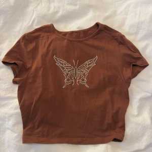 Brun croppad topp med fjärilstryck - Snygg brun croppad topp med korta ärmar och ett glittrigt fjärilsmotiv i strass på bröstet. Perfekt för dig som vill sticka ut med en cool detalj. Mjuk bomullskänsla och rund halsringning.