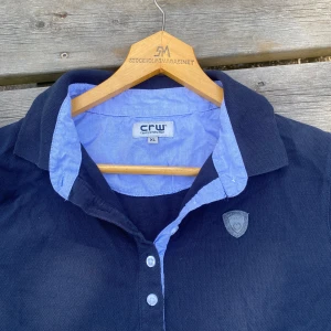 Mörkblå pikétröja från CRW  - Snygg mörkblå pikétröja från CRW i storlek XL med ljusblå krage och knappslå. Tröjan har långa ärmar och ett CRW-märke på bröstet.