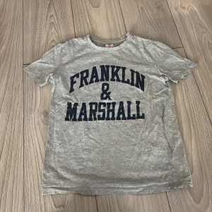 Grå t-shirt Franklin & Marshall - Snygg grå t-shirt från Franklin & Marshall med stor mörkblå logga framtill. Står ingen storlek men skulle säga att det är en Xs/s. Helt oanvänd och ser helt ny ut 