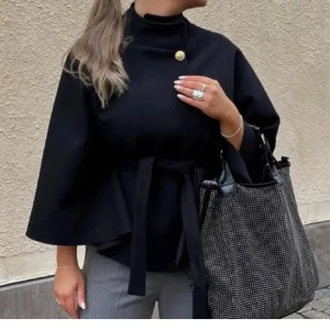 Poncho - Stilren svart poncho med hög krage, vida ärmar och knytbälte i midjan. Snygg detalj med en guldig knapp vid halsen. Perfekt för dig som vill ha en elegant och trendig look.