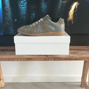 Maison Margiela  - Storlek 46. Kan gå ner till som absolut lägsta 2 799 vid snabb affär. Maison Margiela sneakers i grått med beige detaljer. Ovandel i en mix av mocka och skinn, klassisk gummisula och vita snören. Snygg retrostil med rund tå och diskret branding på sulan. Perfekta för dig som gillar stilrena och tidlösa sneakers. Pris kan diskuteras 