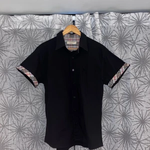 Svart kortärmad Burberry skjorta - Svart kortärmad skjorta från Burberry med klassiskt rutigt mönster på insidan av kragen och ärmsluten. Skjortan har knappar framtill och en stilren, rak passform. Perfekt för dig som gillar en clean look med en touch av lyx.