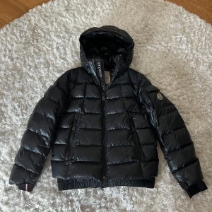 Svart Moncler pufferjacka med huva - Snygg svart pufferjacka från Moncler med huva och dragkedja framtill. Jackan har quiltad design, två fickor med dragkedja och Moncler-logga på ärmen. Perfekt för kalla dagar och riktigt street. Materialet är glansigt och fyllt med dun för extra värme. Osäker på äkthet, dessutom liten i storlek, pass nog s Elr xs//:)