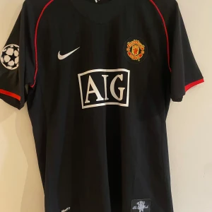 Manchester United Ronaldo 7 Nike tröja - Svart Manchester United fotbollströja från Nike med röda detaljer, AIG sponsortryck och Champions League-märke på ärmen. Ronaldo 7 på ryggen. Tillverkad i lätt Dri-FIT material som andas. Retro tröja från 2008, Aldrig använd