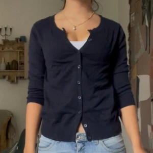 Mörkblå kofta med knappar - Snygg mörkblå kofta med rund hals och långa ärmar. Koftan har en klassisk design med knappar hela vägen framtill och ribbade muddar vid ärmslut. Perfekt att slänga över en t-shirt eller linne för en chill och stilren look.
