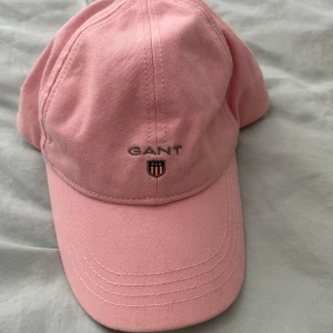 Rosa keps från GANT - Snygg rosa keps från GANT med broderad logga och sköld framtill. Klassisk böjd skärm och justerbar baktill. Perfekt accessoar för en chill och sportig look. Tillverkad i mjuk bomull som känns skön mot huvudet.