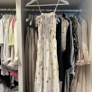 Vit blommig långklänning från H&M - Supersöt långklänning från H&M i vitt med små lila och gula blommor. Klänningen har smala axelband, smock upptill och ett flowigt fall. Perfekt för varma dagar och ger en romantisk vibe. Materialet är mjukt och känns lätt mot huden. Klänningen slutar ungefär vid anklarna. 