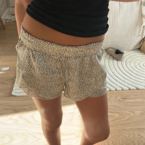 Beige prickiga shorts med volangkant - Säljer ett par beige shorts med svart prickigt mönster och volangkant längs sidorna. De har elastisk midja och är loose fit, perfekta för varma dagar. Materialet känns lätt och luftigt, vilket gör dem sköna att bära under sommaren.