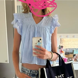 Ljusblå blus med volangärmar - Supersöt ljusblå blus med v-ringning och broderade detaljer upptill. Blusen har korta volangärmar och är croppad i modellen, vilket ger en luftig och trendig look. Perfekt för varma dagar och enkel att styla med jeans eller kjol.