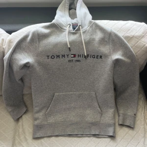 Tommy Hilfiger Hoodie Grå - Snygg grå hoodie från Tommy Hilfiger med broderad logga och text på bröstet. Klassisk känguruficka framtill, vita snören i huvan och ribbade muddar. Perfekt för en chill och stilren look. Ser ut som ny