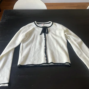 Vit kofta med svarta detaljer från Nelly - Vit kofta från Nelly i stickat material, med svarta kanter, knappar framtill och en rosett vid halsen. Använd en gång.