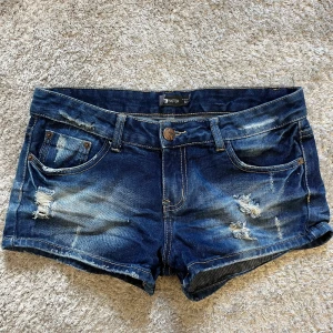 Mörkblå jeansshorts från FB Sister - Snygga mörkblå jeansshorts från FB Sister med slitna detaljer. Shortsens bakficka har en unik dödskallepatch i blått och vitt. Perfekta för en avslappnad och edgy stil i sommar.