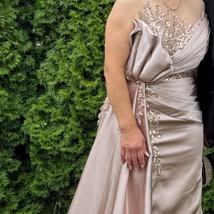 Champagnefärgad långklänning med paljetter - Säljer en elegant långklänning i champagnefärgad satin med glittriga paljettdetaljer och broderier upptill. Klänningen har offshoulder-ärmar, draperad design och vacker volang vid axeln. Perfekt för bal eller fest, ger en lyxig och glamorös känsla.