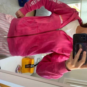 Rosa hoodiejacka från Gymgrossisten - Säljer en rosa hoodiejacka från Gymgrossisten med dragkedja, huva med snörning och tryck på bröstet samt ena ärmen och ryggen. Jackan har kängurufickor och är i mjukt bomullsmaterial. Perfekt för chill eller gymmet.