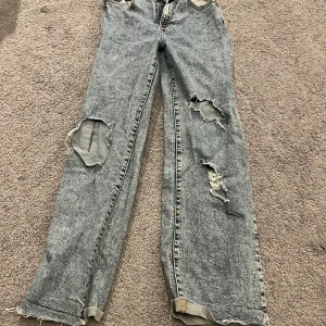 Grå slitna jeans  - Säljer ett par grå, acid wash jeans med raka ben och flera stora slitningar på framsidan. Klassisk femficksmodell. Jeansen har råa kanter vid bensluten för en extra edgy vibe.