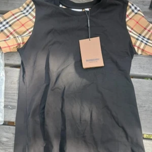 Burberry svart t-shirt med rutiga ärmar - Svart t-shirt från Burberry med klassiska beige och rödrutiga korta ärmar. Snygg kontrast mellan den svarta kroppen och de ikoniska rutmönstrade ärmarna. Rund halsringning och mjukt bomullsmaterial. Perfekt för dig som gillar stilrena designer med en twist.