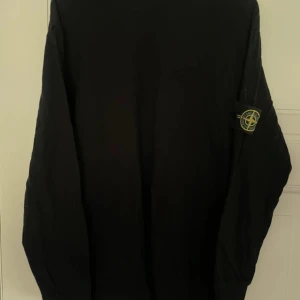 Svart långärmad tröja Stone Island - Svart långärmad tröja från Stone Island med klassisk patch på ärmen. Tröjan har en clean look och är tillverkad i mjuk bomull, perfekt för dig som gillar streetwear och vill ha något stilrent med ikonisk logga.