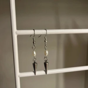 Silvriga örhängen med pärlor och spikes - Snygga hängande örhängen med silvriga spikes och vita pärlor. Perfekt för dig som gillar en edgy stil med en touch av elegans. Örhängena har metallkrokar och cool konformad detalj längst ner. I rostfritt stål och handgjort 