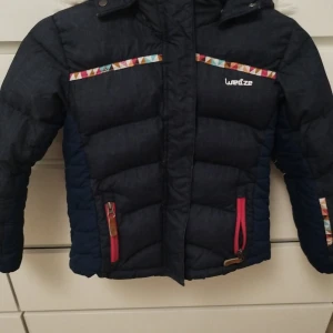 Mörkblå dunjacka för barn - Mörkblå dunjacka med färgglada detaljer och rosa dragkedjor. Jackan har huva med fuskpäls, quiltad design och mönstrade band över bröstet. Insidan är röd och jackan är perfekt för kalla dagar. Snygg och funktionell för vintern! Har några små defekter på armarna men inget som påverkar funktionen av jackan.