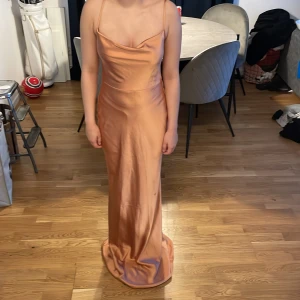 Champagnefärgad långklänning Pretty Lavish - Säljer en elegant långklänning från Pretty Lavish i en lyxig champagnefärg. Klänningen har smala axelband, omlott-detaljer i ryggen och en mjukt draperad urringning. Materialet är glansigt och känns som satin, vilket ger en exklusiv vibe. Perfekt för dig som vill sticka ut med stil.