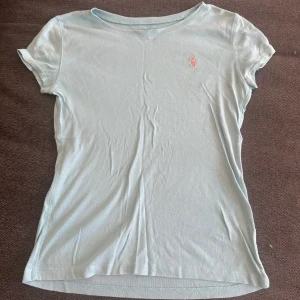 Ljusblå t-shirt från Polo Ralph Lauren - Ljusblå t-shirt från Polo Ralph Lauren med klassisk rund hals och korta ärmar. På bröstet finns den ikoniska rosa broderade loggan. T-shirten är i mjuk bomull och har en enkel, stilren look som passar perfekt till jeans eller shorts.