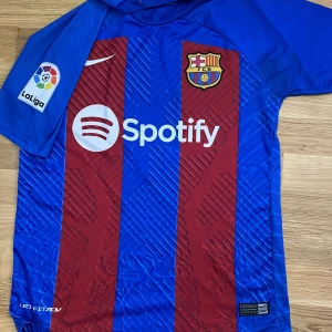 Barcelona Gavi/ spelarversion - Säljer en officiell FC Barcelona matchtröja med Gavi och nummer 6 på ryggen. Tröjan är kortärmad, har breda röda och blå ränder, Barcelona-emblem på bröstet och La Liga-märke på ärmen. Materialet är lätt och andas, perfekt för fotboll. Sponsorloggor från Spotify och UNHCR.
