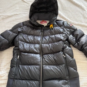 Svart dunjacka från Parajumpers - Snygg svart dunjacka från Parajumpers med huva sytt på armen då den fick ett hål  och därför är priset lägre. Jackan har quiltad design, dragkedja framtill och två sidofickor med dragkedja. Perfekt för kalla dagar och har en cool patch på axeln.